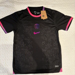 PSG x Nike svart fotbollströja - Snygg svart Paris Saint-Germain fotbollströja från Nike med diskret blommönster i tyget. Tröjan har korta ärmar, rosa och lila detaljer vid krage och ärmslut samt ett unikt rosa Nike-märke på bröstet. Tillverkad i lätt och ventilerande polyester.