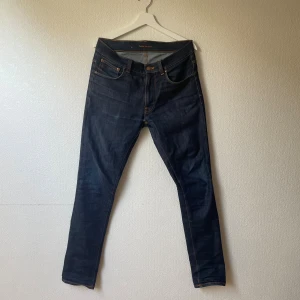 selvedge Nudie Jeans - Snygga mörkblå jeans från Nudie Jeans med klassisk orange söm och vågig detalj på bakfickorna. Modellen har raka ben och normal passform. Jeansen är tillverkade i slitstarkt bomullsjeansmaterial och har en klassisk femficksdesign. 32/34