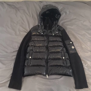 Svart dunjacka med stickade ärmar från Moncler - Snygg svart dunjacka från Moncler med glansig pufferfront och huva. Ärmarna är ribbstickade för en cool kontrast och jackan har dragkedja framtill samt logga på ärmen. Allt funkar NFC scanner allt. Den e i storlek L men passar även för M. Perfekt för dig som vill ha en jacka med både stil och som håller dej varm. Pris ej hugget i sten. 