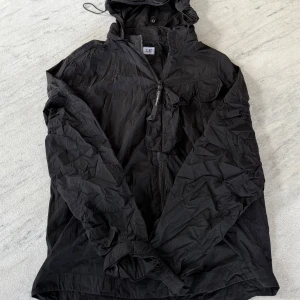 Svart vindjacka från C.P. Company - Svart vindjacka från C.P. Company, modell Goggle Jacket Chrome-R. Jackan har huva, dragkedja framtill, flera fickor med lock och tryckknappar samt detaljerad logotyp på dragkedjan. Tillverkad i återvunnen nylon med lätt, sportig känsla.