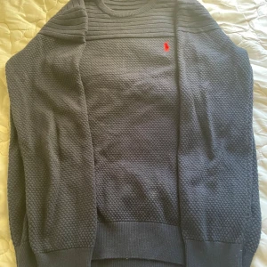 Mörkblå stickad tröja från Polo Ralph Lauren - Säljer en mörkblå stickad tröja från Polo Ralph Lauren med röd broderad logga på bröstet. Tröjan har ribbade detaljer upptill och strukturstickat mönster på resten av plagget. Rund halsringning och långa ärmar. Perfekt för kyligare dagar. Ser lite brun ut men det var ljuset när jag tog bilderna.