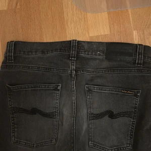 Svarta jeans från Nudie Jeans, W31 L30 - Säljer ett par svarta jeans från Nudie Jeans med klassisk femficksdesign och broderade bakfickor. Jeansen har en smal passform och är tillverkade i mjukt denimtyg. Perfekta för dig som gillar stilrena och bekväma jeans.