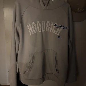 Grå hoodie från Hoodrich strl L - Säljer en stilren grå hoodie från Hoodrich i storlek L. Tröjan har broderad logga framtill, textdetaljer på ryggen och en stor känguruficka. Mjuk insida och huva med broderade detaljer. Perfekt för dig som gillar streetwear och snygga basics.