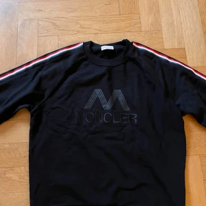 Svart Moncler sweatshirt med ränder - Svart sweatshirt från Moncler med broderad logga framtill och klassiska Moncler-ränder i rött, vitt och blått längs ärmarna. Tröjan har rund halsringning och långa ärmar. Perfekt för dig som gillar stilrena och sportiga vibes.   