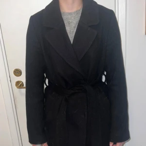 Svart kappa från H&M - Snygg svart kappa från H&M med knytskärp i midjan. Perfekt för kyligare dagar med sin klassiska design och långa ärmar. Ett stilrent val för garderoben.