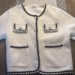 Vit cardigan jacka från Giracoo - Superfin vit cardigan jacka från Giracoo med svart och vit tvinnad kant, dekorativa knappar (saknar en knapp men det märks ej av mycket) och fyra fickor framtill. Tröjan har en fluffig textur och rund halsringning. 