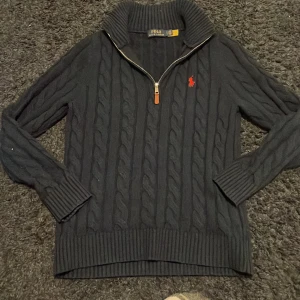 Svart kabelstickad tröja Polo Ralph Lauren - Kabelstickad svart tröja från Polo Ralph Lauren med halv dragkedja och hög krage. Klassisk röd logga broderad på bröstet. Tillverkad i en mix av ull, akryl och nylon. Långärmad och ribbade muddar för extra stilpoäng.