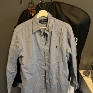 Blå skjorta från Ralph Lauren - Klassisk ljusblå skjorta från Ralph Lauren med button-down krage och broderad logga på bröstet. Skjortan har lång ärm, normal passform och är tillverkad i mjuk bomull. Perfekt för en clean och stilren look.