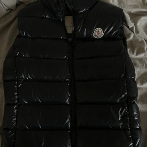 Svart dunväst från Moncler - Säljer en svart, glansig dunväst från Moncler med klassisk quiltad design och dragkedja framtill. Västen har Moncler-logga på bröstet och coolt serietidningsfoder på insidan. Perfekt för lager-på-lager och snygg till streetwear. Kvitto osv följer med. Storlek 1 motsvarar S. 