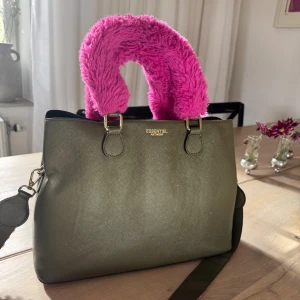 Grön handväska från Essentiel Antwerp - Grön handväska från Essentiel Antwerp med fluffigt rosa handtag och bred axelrem med mönster i rosa, lila och grönt. Väskan är i skinnimitation och har guldfärgade detaljer. 