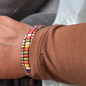 Färgglatt pärlarmband med knäppning - Unikt armband med pärlor i regnbågens färger, inslag av silverfärgade detaljer och brunt snöre. Armbandet har en knapp som knäppning och ger en cool, bohemisk vibe. Perfekt accessoar för att liva upp din stil.