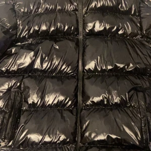 Moncler jacka L - Snygg svart pufferjacka från Moncler i storlek L. Jackan har glansig finish, Moncler-logga på ärmen och dragkedja framtill. Fylld med dun och perfekt för kalla dagar. Klassisk design med quiltade paneler och praktiska fickor.