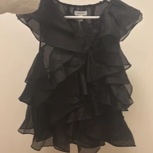 Svart volangblus från H&M Edition - SLUTSÅLD Supersnygg svart blus från H&M Edition med flera lager av volanger och transparent tyg. Blusen är ärmlös och har en luftig, flowy känsla som ger en cool och trendig look. Perfekt för dig som vill sticka ut med en statement piece.