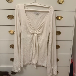 Vit långärmad topp med knyt fram - Snygg vit långärmad topp från design by si med djup v-ringning och knyt fram som ger en feminin touch. Toppens material känns mjukt och lätt, perfekt för lager-på-lager. Enkel och stilren design som passar till många olika outfits.