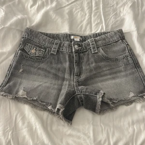 Grå jeansshorts från Gina Tricot - Snygga grå jeansshorts från Gina Tricot med slitna detaljer och råa kanter. Shortsen har broderade bakfickor med paisleymönster och klassisk femficksdesign. Perfekta för en chill sommarlook.