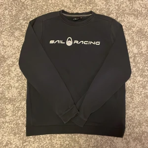 Svart Sail Racing sweatshirt - Svart sweatshirt från Sail Racing med vit logga framtill och liten logga i nacken. Klassisk rund halsringning och långa ärmar. Tröjan har en clean look och är tillverkad i mjukt material som känns skönt mot huden.