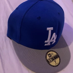 Blå LA Dodgers keps New Era 59FIFTY - Snygg blå LA Dodgers keps från New Era 59FIFTY med grå skärm och broderad LA-logga framtill. Kepsen har klassisk passform, MLB-logga bak och New Era-logga på sidan. Tillverkad i bomull och polyester för en bekväm känsla.
