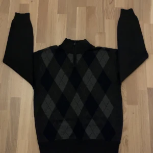 Svart argyletröja med half zip Dylan Star - Snygg svart stickad tröja från Dylan Star med klassiskt argylemönster i grått och mörkgrått framtill. Tröjan har ribbad krage och en praktisk half zip-dragkedja. Perfekt för dig som gillar stilren och tidlös design.