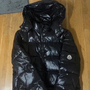 Svart Moncler pufferjacka  - Snygg svart pufferjacka från Moncler med glansig finish och avtagbar huva. Jackan har dragkedja framtill, ficka med Moncler-logga på ärmen och klassisk quiltad design. Perfekt för kalla dagar och riktigt street. Storlek M men passar Perfekt för S. Mycket Bra skick, som ny, använd 1 gång. NFC SCAN FUNGERAR!