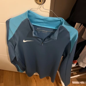 Blå Nike träningströja med half zip - Nike träningströja i blått med ljusblå detaljer på axlar och ärmar. Långärmad modell med half zip framtill och klassisk vit Nike-logga på bröstet. Perfekt för träning eller sportiga dagar. Materialet är mjukt och följsamt syntet.