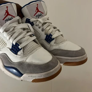 Nike SB Air Jordan 4 White/Blue - Nike SB Air Jordan 4 sneakers i vitt och blått med grå mockadetaljer och gummisula. Skorna har klassisk snörning, meshpaneler och tydlig Jumpman-logga på plösen samt Nike SB branding på hälen. Kombination av skinn och mocka ger en snygg kontrast.