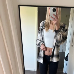 Rutig jacka i beige och svart - Snygg jacka i rutigt mönster med beige, svart och vita toner. Skjortan har knappar framtill, bröstficka och klassisk krage. Perfekt att slänga över en crop top för en vardaglig look men funkar även till fest.. Materialet känns mjukt och lite tjockare, typ bomull eller polyester.