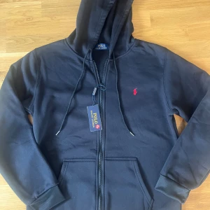 Svart hoodie från Polo Ralph Lauren - Svart hoodie från Polo Ralph Lauren med dragkedja, huva och snörning. Klassisk röd logga broderad på bröstet och två fickor framtill. Tillverkad i mjuk bomullsmix, perfekt för höst och vinter. Snygg och enkel stil som funkar till allt.
