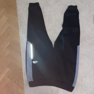 Svarta mjukisbyxor från The North Face - Svarta mjukisbyxor från The North Face med gråa paneler längs sidorna och reflexdetalj på ena benet. Byxorna har resår i midjan och vid benslut, samt logga tryckt på låret. Perfekta för chill eller träning.