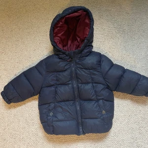 Mörkblå pufferjacka från Zara BabyBoy - Mysig mörkblå pufferjacka från Zara BabyBoy med huva och vinrött foder. Jackan har dragkedja framtill, två fickor med knapp och är perfekt för kyliga dagar. Passar barn i storlek 86, 12-18 månader.