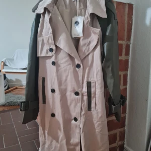 Beige och grön trenchcoat från New Yorker - Snygg trenchcoat i beige med kontrasterande gröna ärmar och detaljer. Jackan har dubbelknäppning, stora slag och bälte vid ärmsluten. Materialmix med bomullskänsla i kroppen och skinnimitation på ärmarna. Perfekt för dig som vill sticka ut med en trendig look.