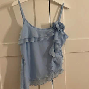 Ljusblå volangtop från SHEIN - Supersöt ljusblå linnetopp från SHEIN med tunna axelband och volangdetaljer framtill. Toppen har en dekorativ blomma på ena axelbandet och asymmetrisk nederkant. Perfekt för dig som gillar romantiska och lekfulla plagg.
