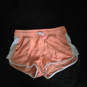 Persikofärgade shorts från H&M - Snygga persikofärgade shorts från H&M med vit kant och elastisk midja med snörning. De har en sportig look och är perfekta för varma dagar. Materialet är mjukt och bekvämt, troligen bomull. Passformen är normal och modellen är kort.