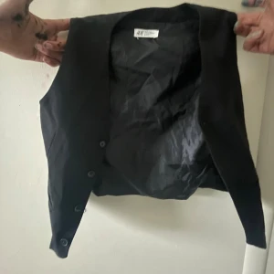 Svart kostymväst från H&M - Klassisk svart väst från H&M med knäppning framtill och justerbart spänne i ryggen. Snyggt figursydd modell i slät polyester, perfekt att styla över skjorta eller t-shirt för en clean look.