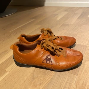 Bruna sneakers från Le Coq Sportif - Snygga bruna sneakers från Le Coq Sportif i skinn med klassisk snörning och svart logga på sidan. Skorna har rund tå, platt svart sula och en stilren design som passar till det mesta. Perfekta för dig som gillar retrovibbar och fransk sportstil. Pris kan diskuteras!