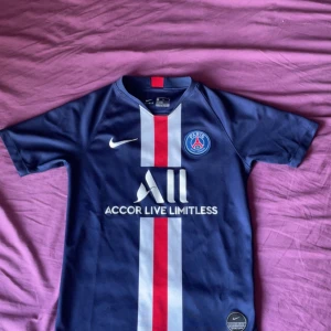 PSG Tröja från Nike, STL 137-147 CM - Paris Saint-Germain fotbollströja från Nike i Dri-FIT-material. Marinblå med vit och röd vertikal rand, PSG-emblem på bröstet och sponsortryck. Klassisk kortärmad modell, perfekt för dig som älskar fotboll och vill bära klubbens färger. 