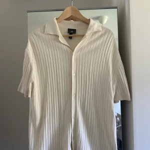 Ribbad vit skjorta från H&M - Säljer en ribbad vit skjorta från H&M med korta ärmar och klassisk krage. Skjortan har knappar hela vägen och är i ett mjukt, stickat material som ger en chill och modern vibe. M