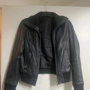 svart skinnbomberjacka - Snygg svart bomberjacka i skinn med ribbade muddar och krage. Jackan har dragkedja framtill och två fickor med dragkedja. Perfekt för dig som vill ha en stilren och edgy look.