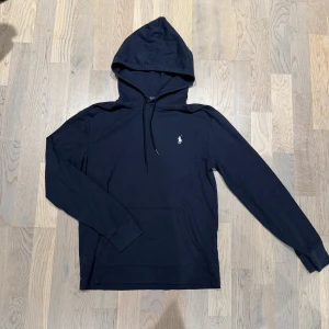 Mörkblå Ralph Lauren hoodie - Svart hoodie från Polo Ralph Lauren i bomull med klassisk huva och dragsko. Liten broderad logga i vitt på bröstet och känguruficka framtill. Perfekt för en chill och stilren look.