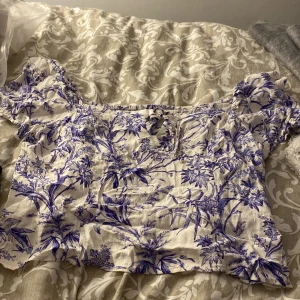 Blommig offshoulder blus i blå och vit - Supersöt blus med offshoulder och korta puffärmar. Topp i vit bas med blått blommigt mönster, smockad rygg och liten knytning framtill. Luftigt material, perfekt för varma dagar. Storlek L men skulle säga M då den krympt 
