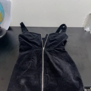 Svart manchesterklänning med dragkedja - Snygg svart klänning i manchester med smala axelband och en markerad dragkedja framtill. Klänningen har en figurnära passform och är perfekt för dig som gillar en stilren look med lite edge.