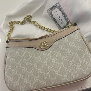 Beige axelväska från Gucci med kedja - Säljer en snygg beige axelväska från Gucci med klassiskt GG-mönster och guldfärgad kedja. Väskan har dragkedja upptill och detaljer i skinn. Perfekt storlek för det viktigaste och en riktigt trendig accessoar. 