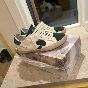The Gallerist vita och gröna sneakers - Snygga sneakers från The Gallerist med vit skinnbas och gröna mockadetaljer på tå och häl. Sidan pryds av en stor grön klöver, och hälen har grönt parti med text. Klassisk snörning och platt sula. Kommer med originalkartong med konstnärligt motiv. 
