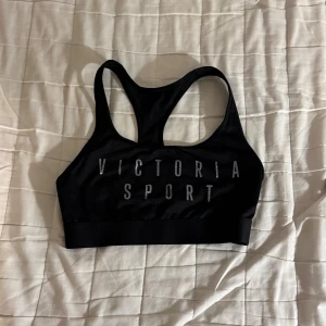 Svart sporttopp Victoria Sport S - Svart sporttopp från Victoria Sport med bred resår och racerback. Toppen har tryckt logga framtill i silver och är tillverkad i ett stretchigt syntetmaterial som passar perfekt för träning.