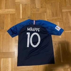 Frankrike Mbappé 10 Nike matchtröja - Säljer en officiell Frankrike fotbollströja från Nike med Mbappé och nummer 10 på ryggen. Tröjan är mörkblå med blå ärmar, har FIFA-märke på bröstet och franska landslagets emblem. Tillverkad i lätt och ventilerande material, perfekt för fotbollsfans.