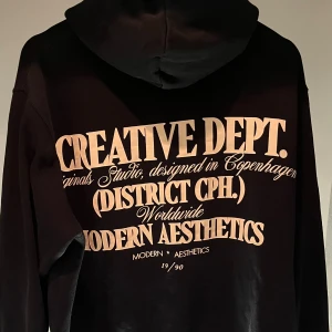 Svart hoodie från Jack & Jones - Svart hoodie från Jack & Jones med stor beige text på ryggen och mindre tryck på bröstet. Klassisk känguruficka framtill och huva. Perfekt för dig som gillar streetstyle och tydliga prints. Inga defekter!! 