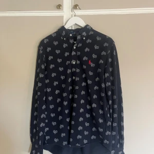 Mörkblå skjorta från Ralph Lauren - Snygg mörkblå skjorta från Ralph Lauren med coolt cykelmönster i vitt och klassisk röd broderad logga på bröstet. Skjortan har knappar framtill, lång ärm och är gjord i lätt bomullsmaterial. Perfekt för dig som gillar unika detaljer och stilren design.