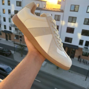 maison margiela - Snygga maison margiela i beige och vitt med ovandel i skinn och mocka, samt klassisk gummisula i brunt. Skorna har rund tå, platt sula och snörning. Perfekta för dig som gillar stilrena och minimalistiska sneakers med retrovibbar.