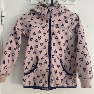 Rosa hjärtmönstrad jacka från H&M - Söt rosa jacka från H&M med mörkblå hjärtan över hela tyget. Jackan har huva, dragkedja framtill och två fickor. Insidan är fodrad med mjuk fleece för extra komfort. Perfekt för kyliga dagar och har ribbade muddar vid ärmsluten.