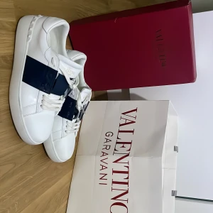 Valentino Garavani Open sneakers vit/blå - Snygga Valentino Garavani Open sneakers i vitt skinn med bred marinblå rem över mitten. Klassisk rund tå, vita snören och diskret logga på plösen. Kommer med originalkartong och påse. Perfekta för dig som vill ha en clean och lyxig look.