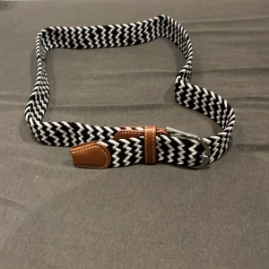 Svartvitt flätat bälte med skinn-detaljer - Snyggt svartvitt flätat bälte med bruna skinn-detaljer och silverfärgat spänne. Bältet har ett coolt zigzag-mönster och passar perfekt till jeans eller chinos. Flexibelt material som gör det bekvämt att bära hela dagen. Helt ny och oanvänd